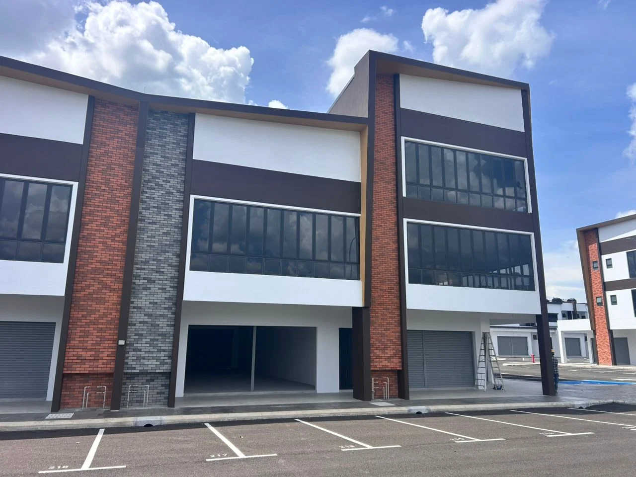 Exterior type: linked terrace house; features: brown and white painted facade, large glass windows, brick accent wall, flat roof, two-storey structure, open parking area, roller shutter doors, concrete walkway, surrounding parking spaces; style: modern.